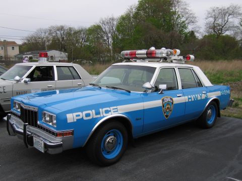 1991 Ford LTD Crown Victoria - blue NYPD highway-patrol Dodge sedan lettered 'POLICE' with a red roof lightbar and blue wheels parked in a lot.