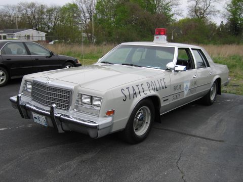 1991 Ford LTD Crown Victoria - silver Chrysler four-door sedan lettered 'STATE POLICE' with a single roof beacon, parked in a lot beside other cars.