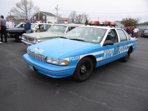 1991 Ford LTD Crown Victoria - blue-and-white NYPD Chevrolet Caprice police sedan with roof beacons parked among classic cars at an outdoor show.