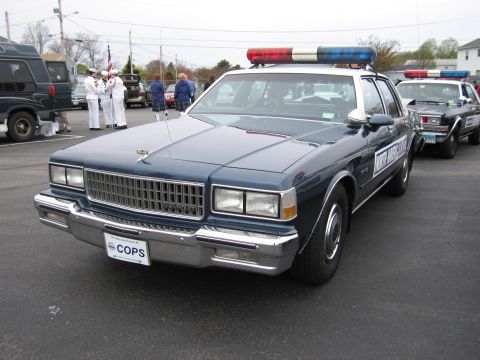 1991 Ford LTD Crown Victoria - dark-blue Chevrolet Caprice police sedan with a red-and-blue roof lightbar parked near a color guard at an event.