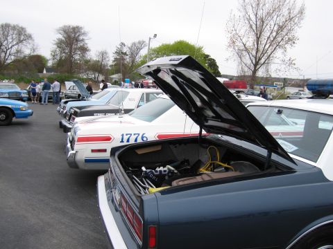1991 Ford LTD Crown Victoria - row of vintage police cars parked in a lot, including a dark sedan with open trunk and a white car lettered '1776'.