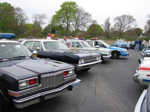 1991 Ford LTD Crown Victoria - diagonal lineup of vintage police sedans with chrome bumpers parked in an asphalt lot with people browsing.