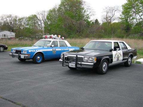 1991 Ford LTD Crown Victoria - blue and white NYPD-marked sedan parks next to a black-and-white Ford police cruiser with a push bar in a paved lot.