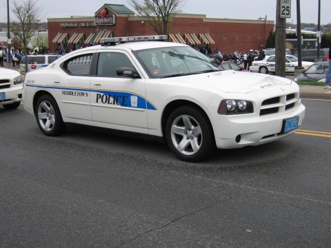 1991 Ford LTD Crown Victoria - white Dodge Charger marked Middletown Police with a blue accent stripe drives along a road past a shopping plaza.