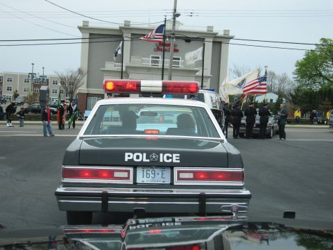 1991 Ford LTD Crown Victoria - rear of a gray police sedan lettered POLICE with red roof lights, parked near American flags and an honor guard.