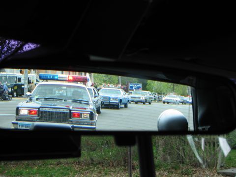 1991 Ford LTD Crown Victoria - line of police cars with lights on approaches, seen through a windshield with the rearview mirror in frame.