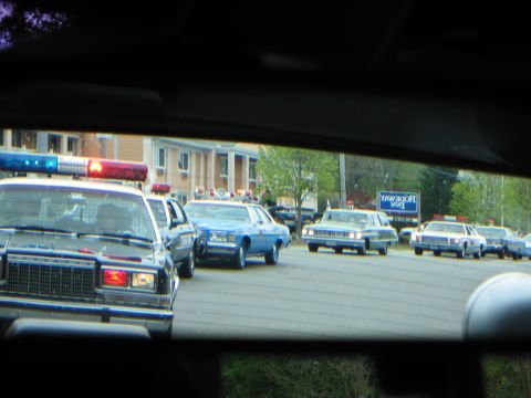1991 Ford LTD Crown Victoria - column of marked police cars with roof lights queued in a lot, viewed through a vehicle's rear window.