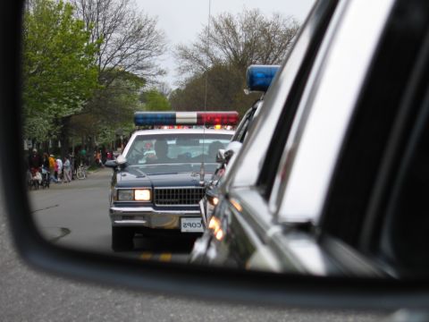 1991 Ford LTD Crown Victoria - black police sedan with red and blue roof lights illuminated, framed in a car's side mirror along a tree-lined street.