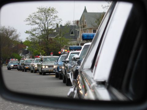 1991 Ford LTD Crown Victoria - long procession of police cars stretches down a residential street, reflected in a vehicle's side mirror.