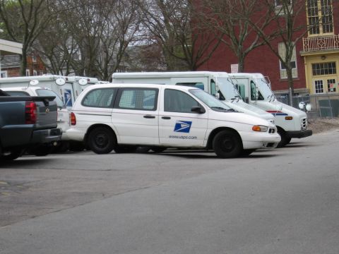 1991 Ford LTD Crown Victoria - white USPS minivan and boxy mail delivery trucks parked beside a pickup near brick buildings.