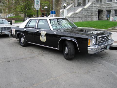 1991 Ford LTD Crown Victoria - black-and-white Ford LTD police sedan with a gold badge decal parked in front of a stone municipal building.