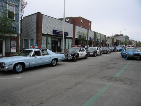 1991 Ford LTD Crown Victoria - lineup of classic police cruisers, led by a light-blue sedan, parked curbside on a downtown street at a car show.