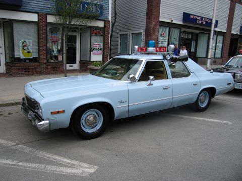 1991 Ford LTD Crown Victoria - restored light-blue Plymouth Fury State Police sedan with a single roof beacon parked along a storefront curb.