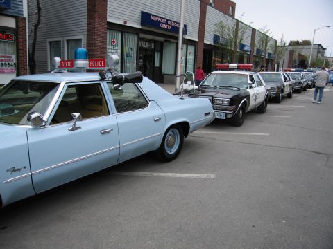 1991 Ford LTD Crown Victoria - light-blue State Police sedan parked in front of a row of black-and-white police cruisers on a downtown street.