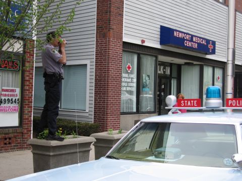 1991 Ford LTD Crown Victoria - uniformed officer stands on the sidewalk beside a light-blue State Police cruiser outside Newport Medical Center.