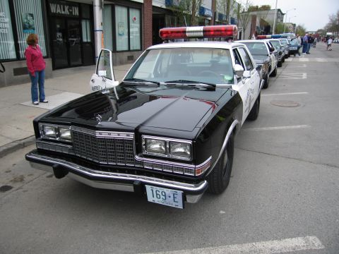 1991 Ford LTD Crown Victoria - black police sedan with white doors, roof lightbar, and plate 169-E parked with its driver door open at a show.
