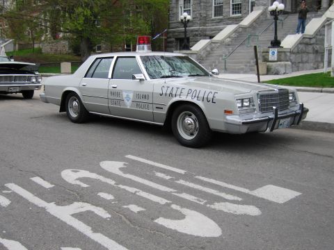 1991 Ford LTD Crown Victoria - silver Chrysler-style Rhode Island State Police sedan with roof beacon parked on a street in front of a stone building.