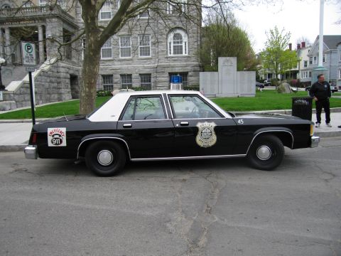 1991 Ford LTD Crown Victoria - black Ford LTD police sedan with white roof and Emergency 911 wheel decal parked beside a stone municipal building.