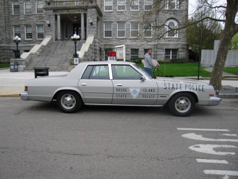 1991 Ford LTD Crown Victoria - Side profile of a silver Rhode Island State Police sedan with roof beacon parked in front of a city hall building.