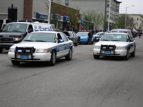 1991 Ford LTD Crown Victoria - Two white-and-blue Ford Crown Victoria police cruisers with light bars rolling down a downtown street in a procession.