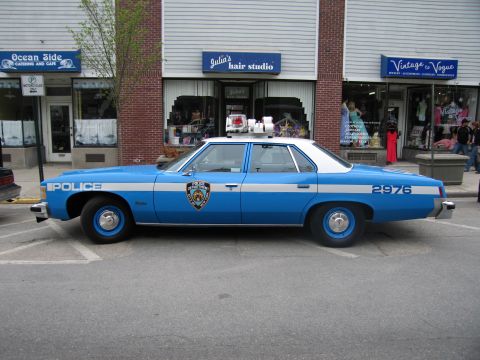 1991 Ford LTD Crown Victoria - Side profile of a blue-and-white NYPD Police sedan, number 2976, parked along a street in front of small shops.