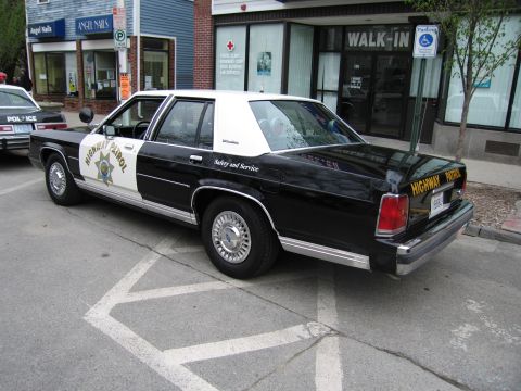 1991 Ford LTD Crown Victoria - Rear three-quarter view of a black-and-white Highway Patrol sedan parked diagonally on a street near a walk-in clinic.