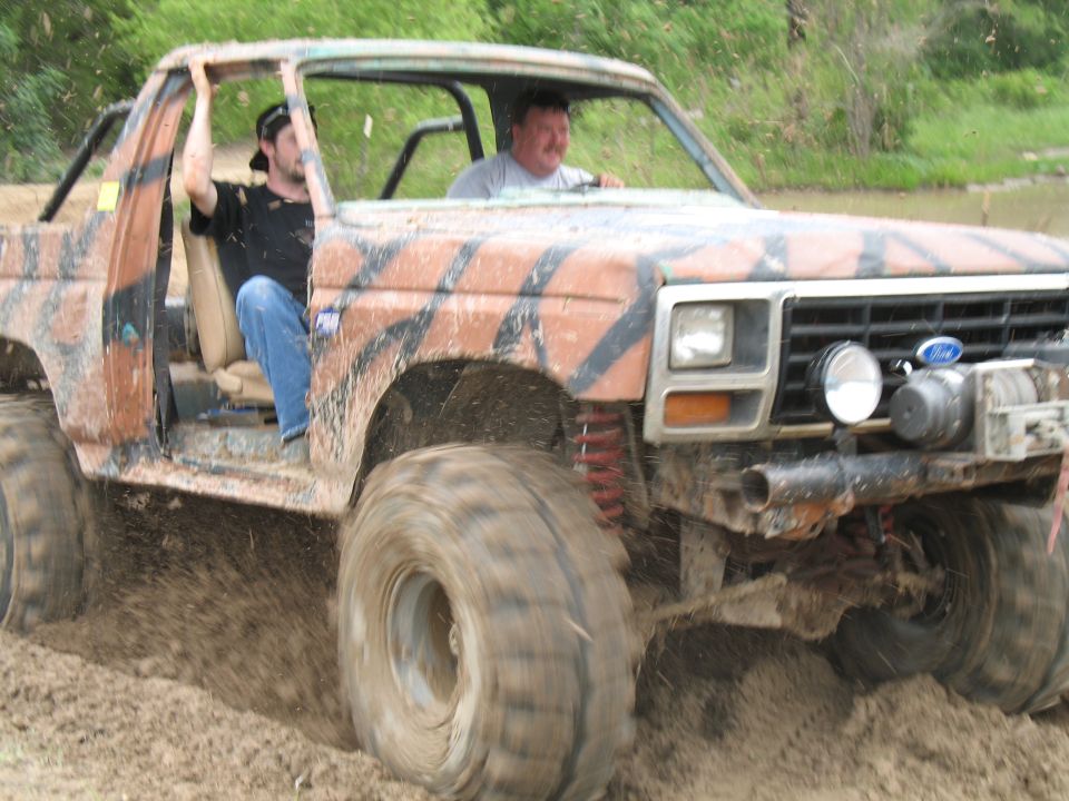 Two men ride in a mud-covered camo Ford with no doors off-road