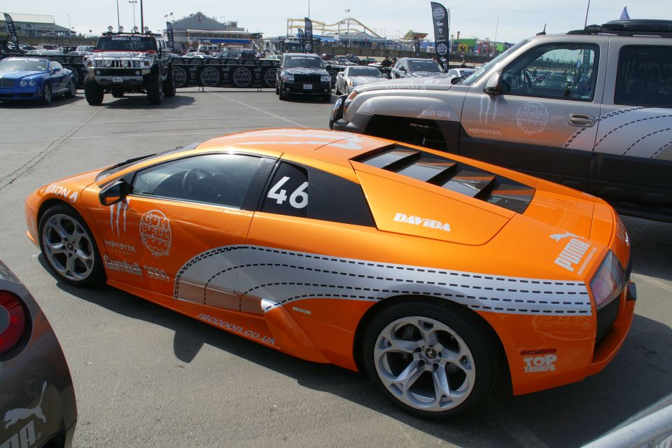 Orange sports car with "GUMBALL 46" and Puma logos, parked next to a silver SUV
