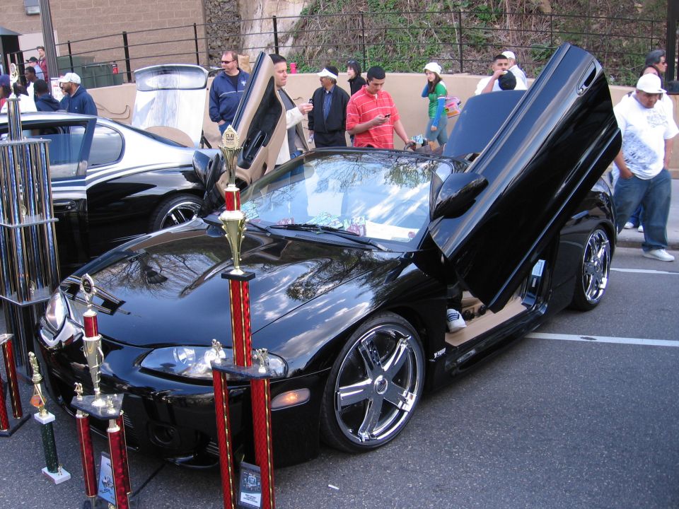 Black coupe with custom lambo doors open, chrome rims, and trophies in front