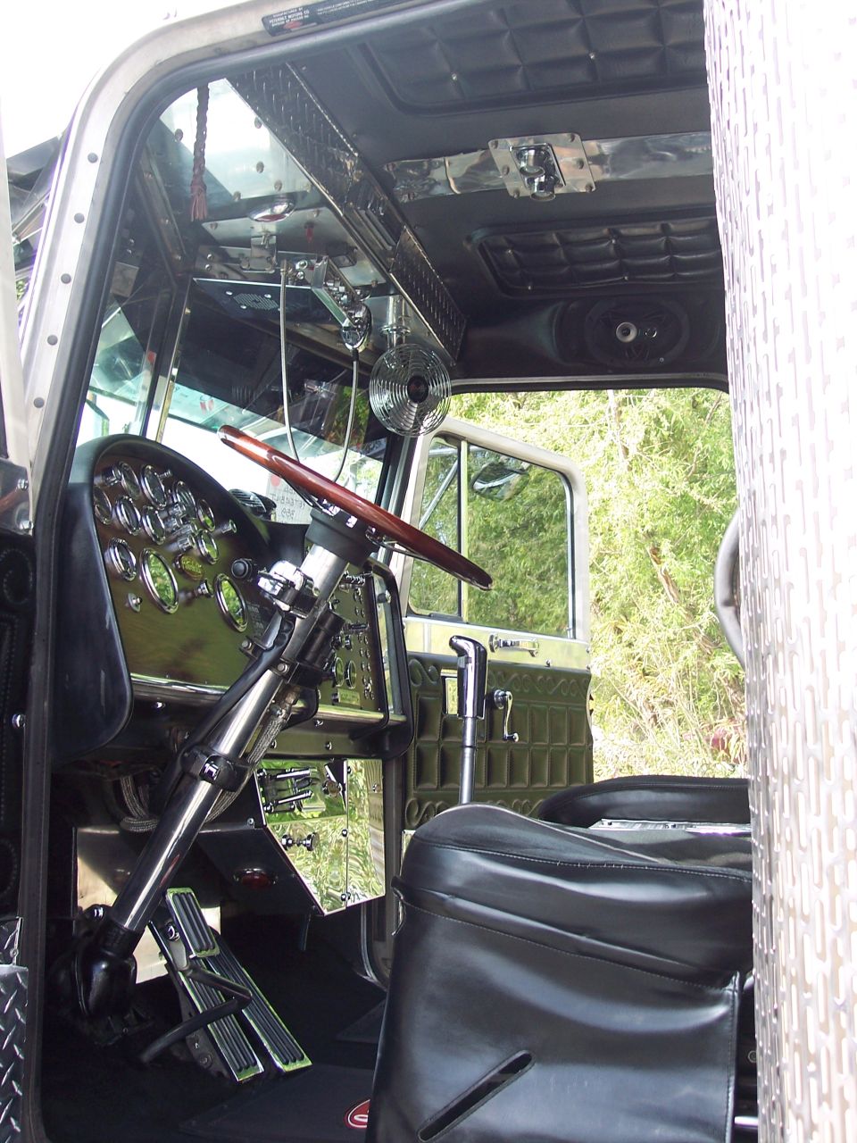 Interior view of a large truck's dashboard, steering wheel, and driver's seat. Chrome accents and gauges are visible