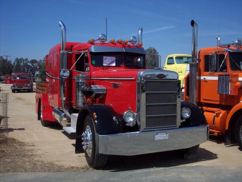 1981 Kenworth W900A - A red Peterbilt 359 semi-truck is parked outdoors with other trucks visible in the background.