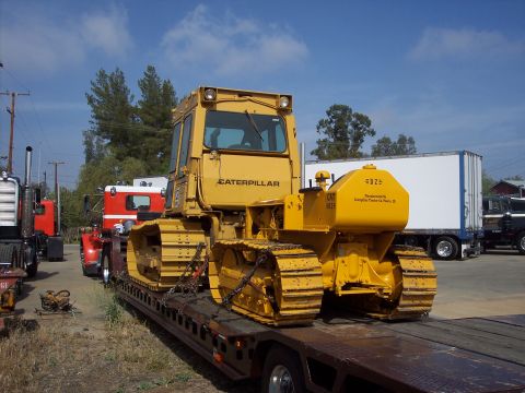 1981 Kenworth W900A - A yellow Caterpillar bulldozer, identified as a CAT 955H, is transported on a flatbed trailer, with other trucks visible in the background.