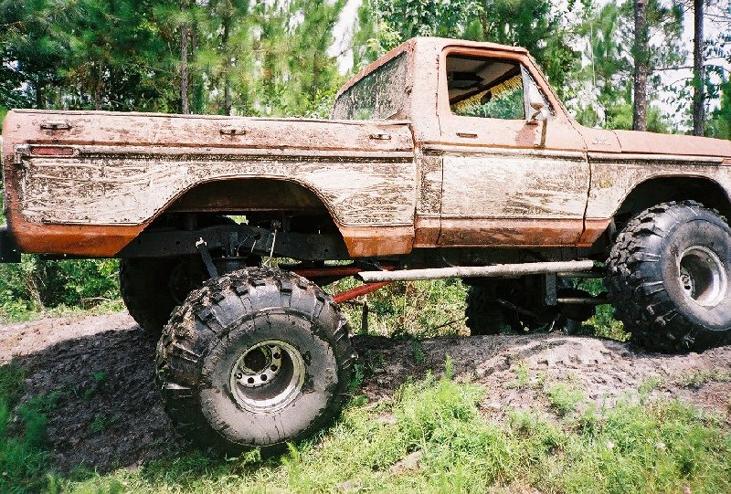 Mud-covered lifted single-cab pickup on tall mud tires parked in a clearing among pine trees