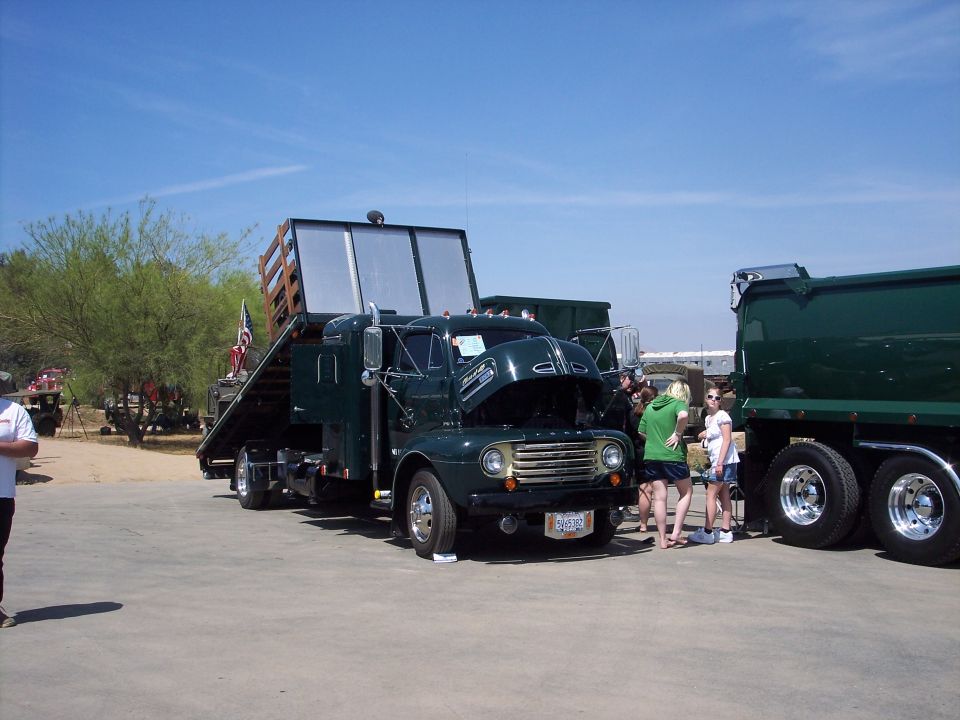 dark green flatbed truck with hood open, trailer up, and a dump truck next to it