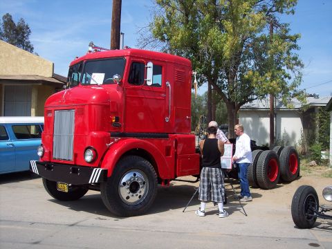 1981 Kenworth W900A - A red semi-truck cab is viewed from a three-quarter front angle, with people and trees in the background.