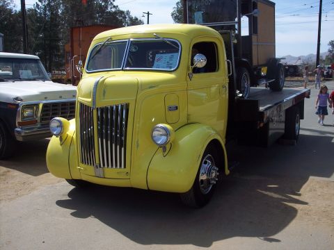1981 Kenworth W900A - A vintage yellow flatbed truck is parked outdoors, with a white pickup truck to its left and people in the background.