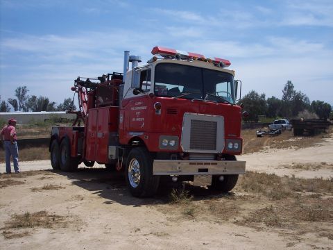 1981 Kenworth W900A - A red tow truck with a white cab is parked on a dirt lot under a blue sky with scattered clouds.