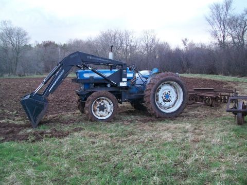 2000 Chevrolet Tahoe - A blue tractor with a front loader is parked in a field with tilled soil and farm equipment nearby.