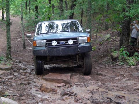 1989 Ford Bronco - A green Ford Bronco navigates a challenging rocky terrain on a wooded trail.