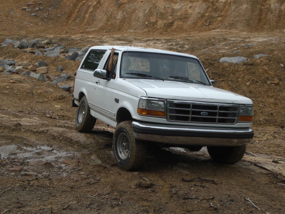 White Ford Bronco driving on a muddy, rocky trail. Driver's arm is out the window