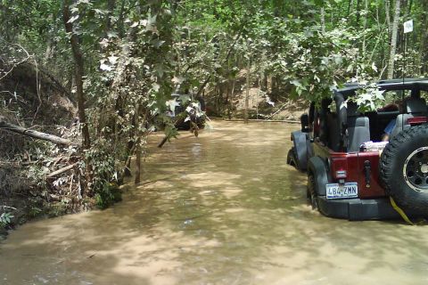 2008 Jeep Wrangler - A red Jeep Wrangler navigates through a muddy, wooded area, with another vehicle visible in the distance.