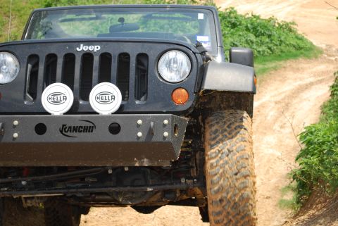2008 Jeep Wrangler - A black Jeep Wrangler is seen from the front, an aftermarket bumper and Hella lights, driving on a dirt road.