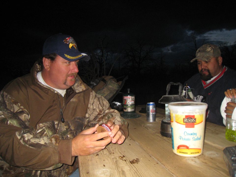 Two men sit at a wooden picnic table outdoors at night. One man holds a small round object. A container of potato salad and a can of soda are on the table