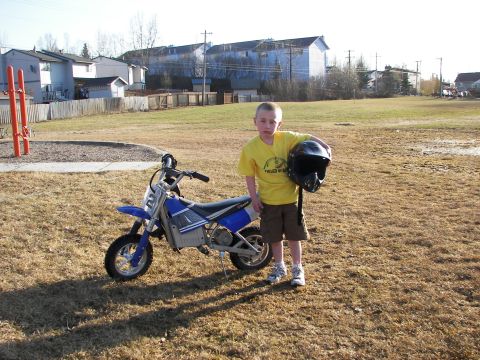 1990 Chevrolet Suburban - A young boy stands with his dirt bike and helmet in a grassy park area.