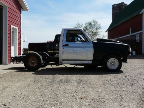 1994 Ford F-150 - This Ford F-series truck chassis is missing its bed and rear fenders, with a black hood and bare metal cab.
