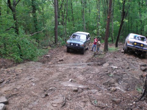 1989 Ford Bronco - Two SUVs are shown navigating a challenging muddy trail in a wooded area.