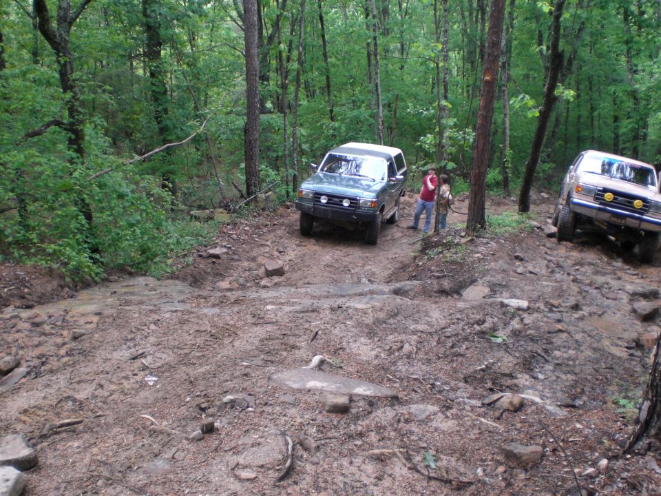 Two SUVs on a muddy forest trail; one is a dark green SUV, the other is a silver SUV with yellow fog lights