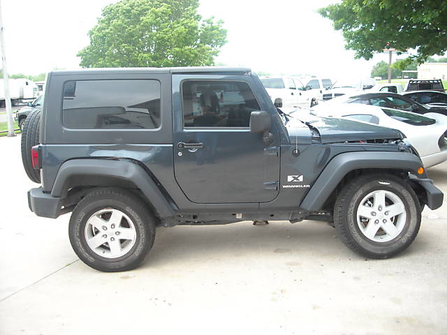 Dark gray Jeep Wrangler parked outside with hood slightly ajar. Other vehicles visible in background