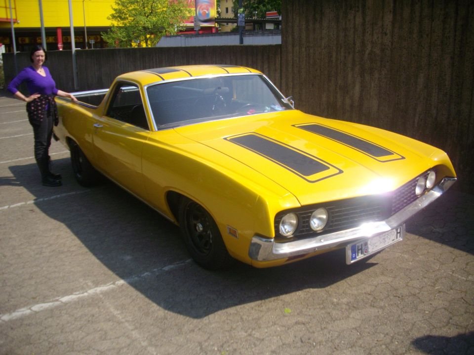 Yellow Ford Ranchero with black racing stripes on the hood and roof. A woman stands next to it