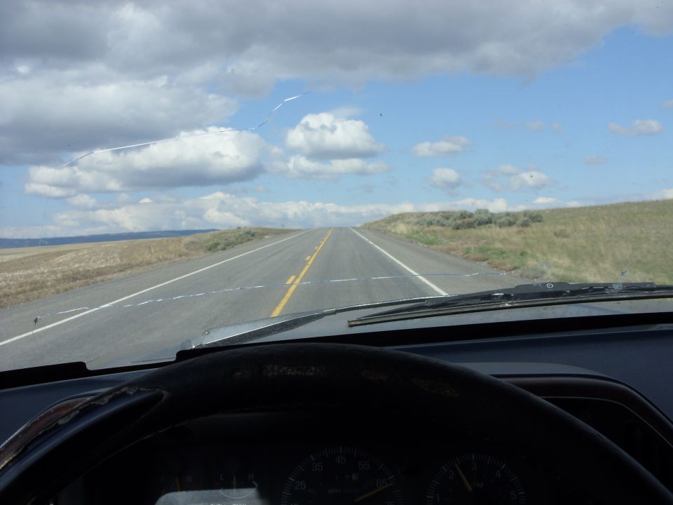 View from inside a vehicle looking out at a long, empty highway stretching to the horizon under a cloudy sky